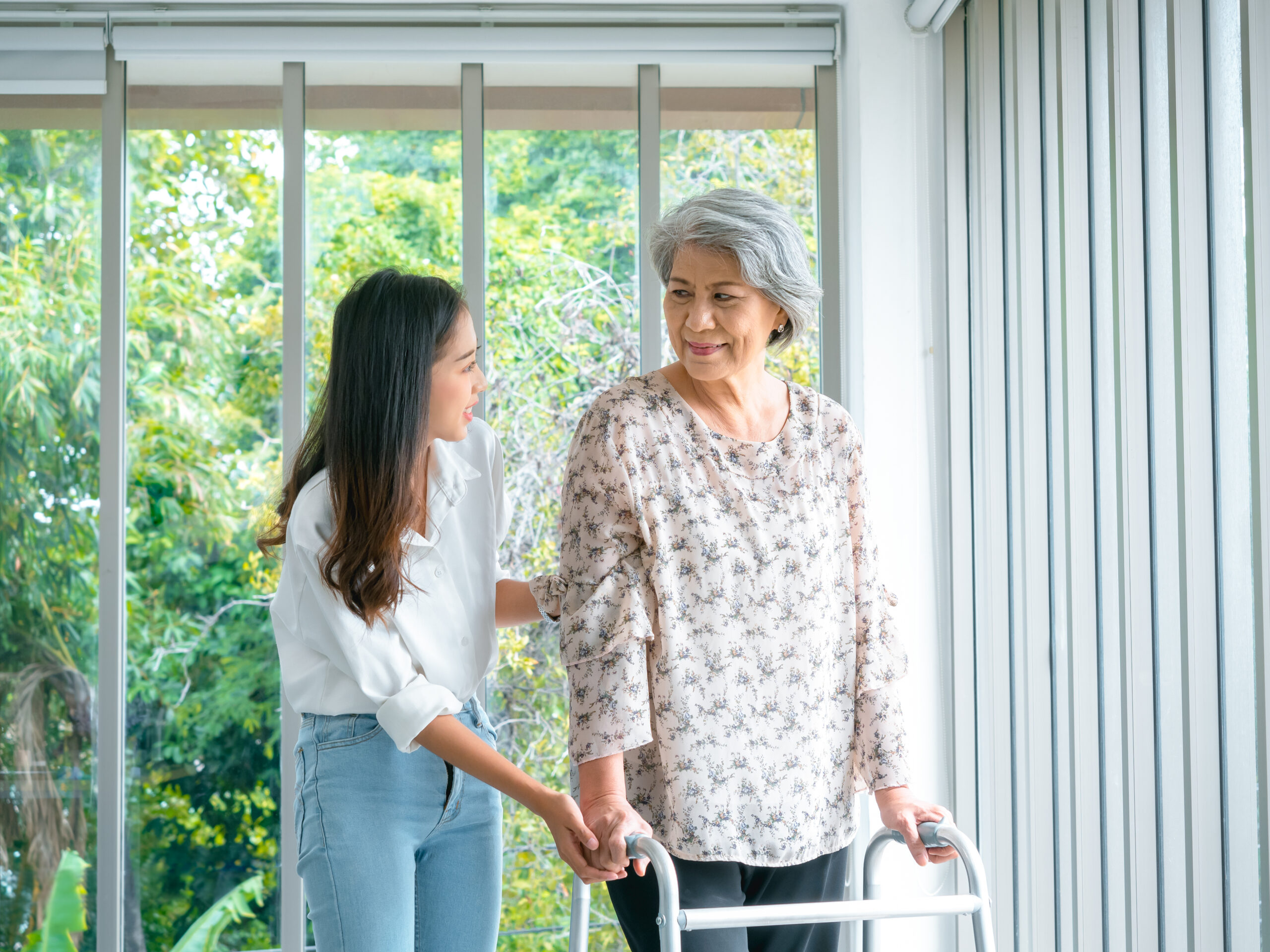 Caregiver, smiling young Asian female, daughter or grandchild supporting help elderly woman, mother or grandparents try to walk with walker frame at home on green nature background, senior healthcare.
